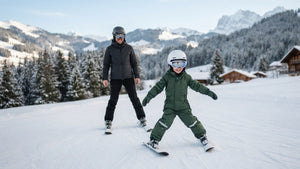 Lächelndes Kind im Skioverall fährt im Schneepflug eine Anfängerpiste hinunter, ein Elternteil fährt hinter ihm