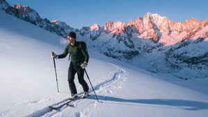 Skitourengeher in grüner erster Schicht aus Merinowolle steigt bei Sonnenaufgang mit Fellen einen verschneiten Hang hinauf, rosa leuchtende Gipfel im Hintergrund