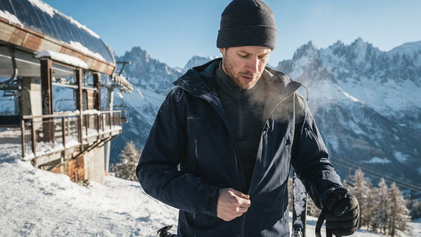 Skifahrer oben am Sessellift, der an einem sonnigen Wintermorgen seine Skijacke über einer ersten Schicht aus Merinowolle mit Reißverschlusskragen schließt